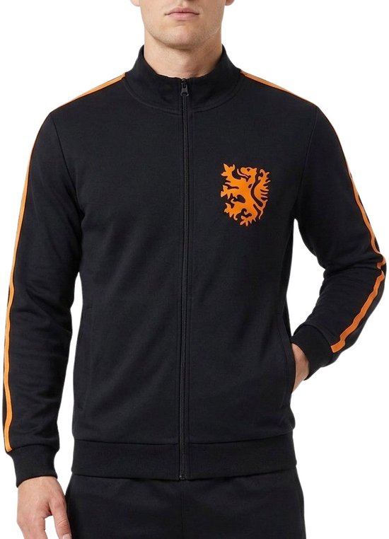Model draagt een zwart Cruyff trainingsjack met oranje strepen op de mouwen en een oranje leeuwenembleem op de borst.