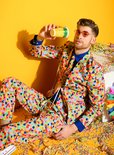 Man met zonnebril en confettibeker in veelkleurig pak met confettiprint, zittend.