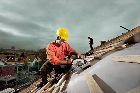 Bouwvakker met gele helm en oranje trui zaagt op dak, met regenachtige lucht.