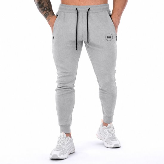 Grijze trainingsbroek met MW logo, ritszakken en trekkoord, gedragen door een man. Getailleerde broek met witte sneakers.
