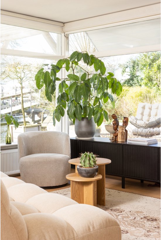 Een lichte fauteuil met een ronde vorm, gestoffeerd in een zandkleurige geweven stof, in een woonkamer bij een raam.