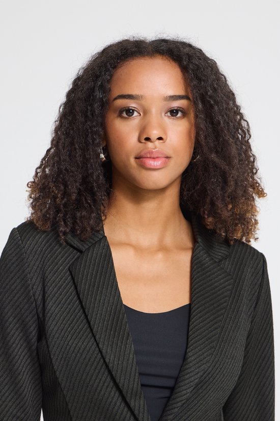 Close-up van een model met krullend haar, draagt een zwarte blazer met diagonaal patroon over een zwart shirt.