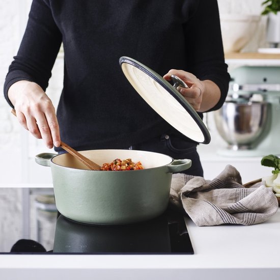 Olijfgroene braadpan met deksel, gevuld met een stoofgerecht, wordt door een persoon geroerd.