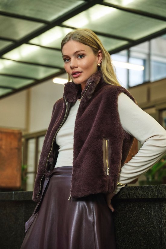 Model draagt een korte bruine faux fur gilet met ritssluiting en gouden details, over een witte top en bruine rok.