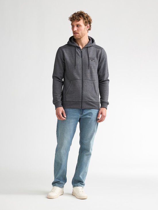 Man met krullend haar draagt een donkergrijs gemêleerd vest met rits en spijkerbroek.