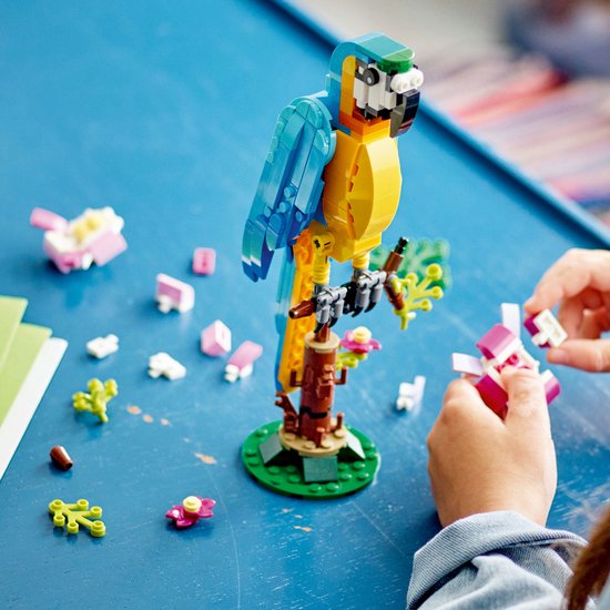 Een gele en blauwe papegaai van LEGO-blokjes op een takje met roze bloemen, omringd door losse blokjes en groene plantjes.