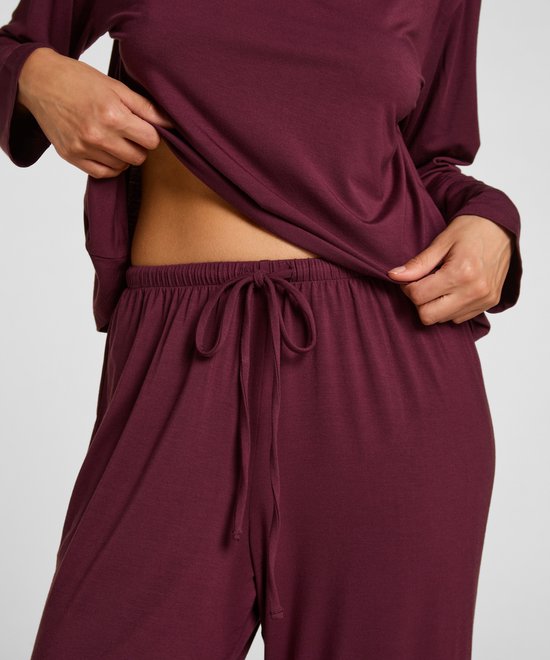 Close-up van een vrouw in een bordeauxrode pyjamaset, met haar handen die de stof van de pyjamabroek met trekkoord optillen.