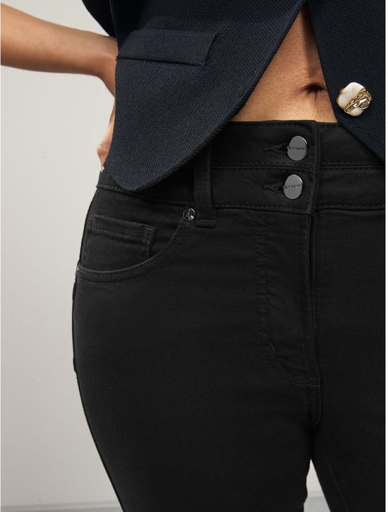 Detail van de tailleband van een zwarte jeans met twee knopen, gedragen onder een donkerblauw vest.