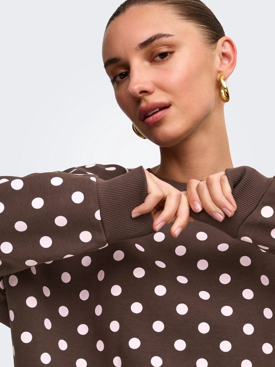 Close-up van een bruine sweater met roze polkadots, gedragen door een model met gouden oorbellen.