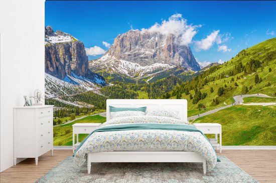 Papier peint représentant un paysage de montagne, des collines verdoyantes, des sommets enneigés, une route sinueuse avec des voitures et une maison en bois. Au premier plan, un lit blanc avec une couette décorative et un tapis.