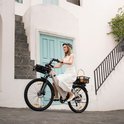 Vrouw met een witte jurk en helm op een champagne gouden elektrische fiets met mandje achterop.