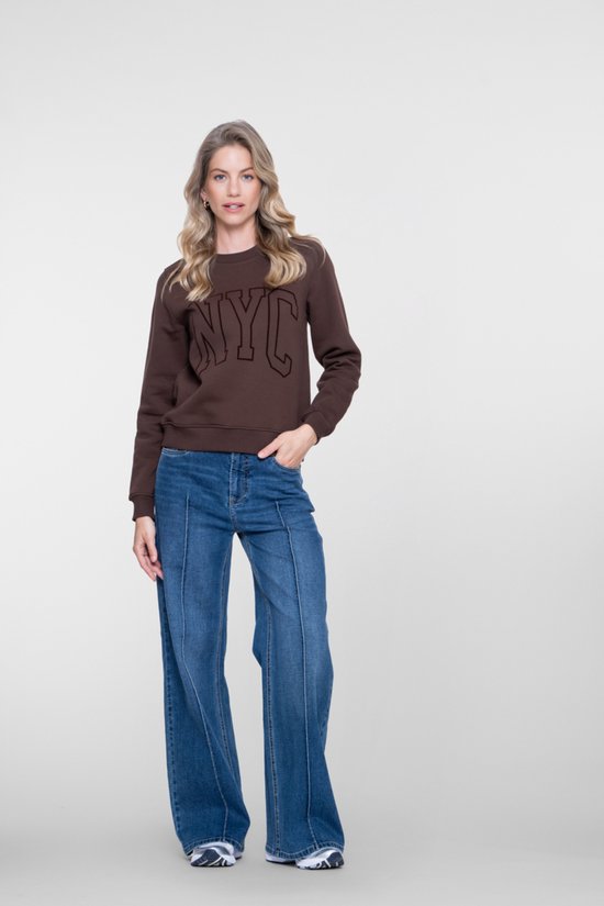 Model met blonde krullen poseert in een bruine 'NYC' sweater en wijde blauwe jeans, kijkend naar de camera.