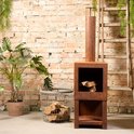 Roestkleurige buitenkachel met schoorsteen, vol met houtblokken, naast een plant en mand met hout.