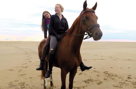 Twee jonge vrouwen rijden op een bruin paard op een strand. Meisje achterop met paars-blauwe jas.