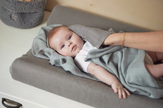 Een baby van 3 maanden ligt op een grijs aankleedkussen, gewikkeld in een grijs muslin doek.