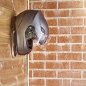 Grijze helm met vizier hangend aan een zilverkleurige haak tegen een bakstenen muur.
