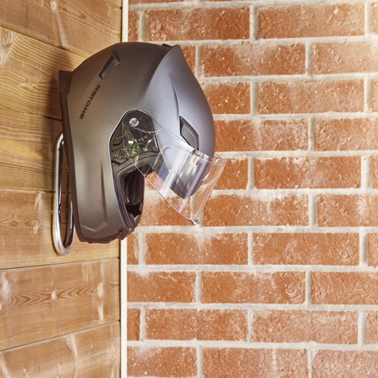 Grijze helm met vizier hangend aan een zilverkleurige haak tegen een bakstenen muur.