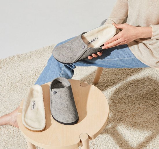 Een persoon met spijkerbroek en beige vest houdt een lichtgrijze vilten Birkenstock pantoffel vast. Een andere pantoffel staat op een krukje.
