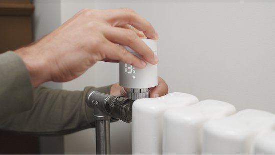 Handen draaien aan een witte radiatorthermostaat met digitaal display op een witte radiator.