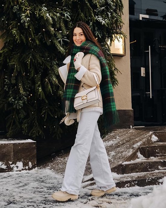 Vrouw met groene en bruine ruitjessjaal om, beige jas en witte broek.
