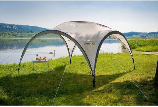 Een witte Coleman partytent staat bij een meer met een tafel met drankjes.