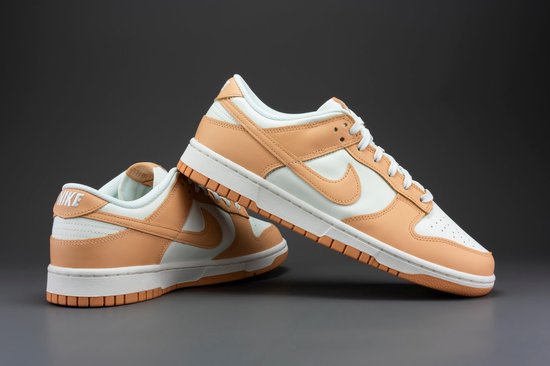 Een paar Nike Dunk Low damesschoenen, wit met oranje overlays, tegen een grijze achtergrond.