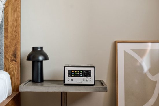 Compacte zwarte radio met zilveren bedieningspaneel staat op een nachtkastje naast een zwarte lamp.