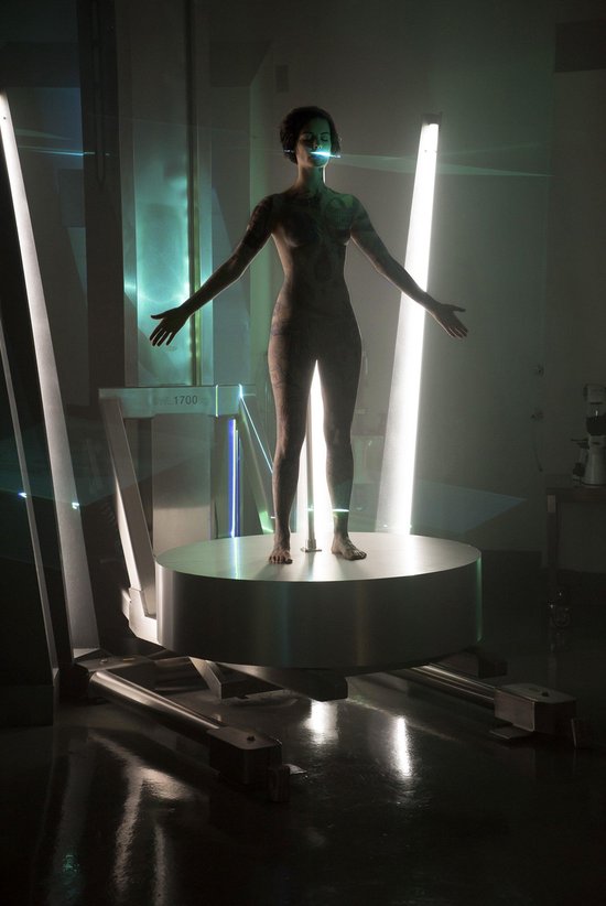 Vrouw met tatoeages op een rond platform in een futuristische kamer met lampen.