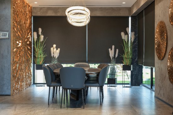 Eetkamer met een grote tafel, grijze stoelen, verduisterende rolgordijnen en gouden wanddecoratie.