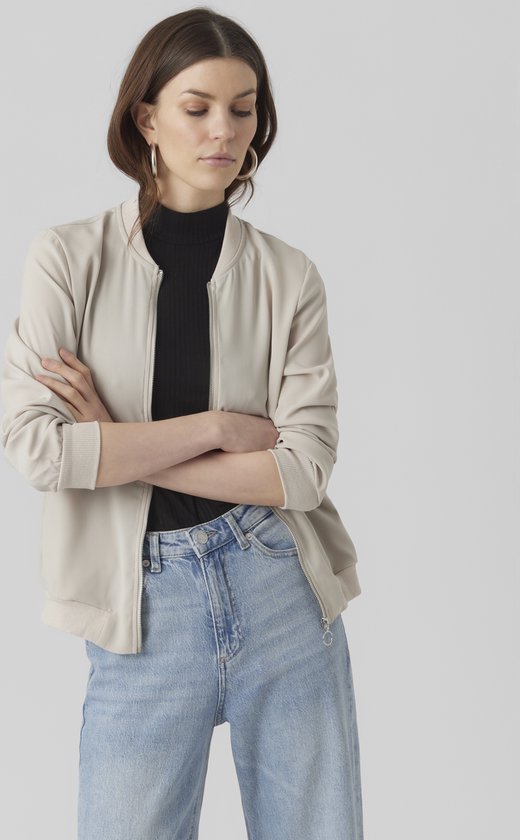 Een model met gekruiste armen, draagt een beige bomberjack over een zwarte coltrui en een spijkerbroek.