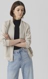 Een model met gekruiste armen, draagt een beige bomberjack over een zwarte coltrui en een spijkerbroek.