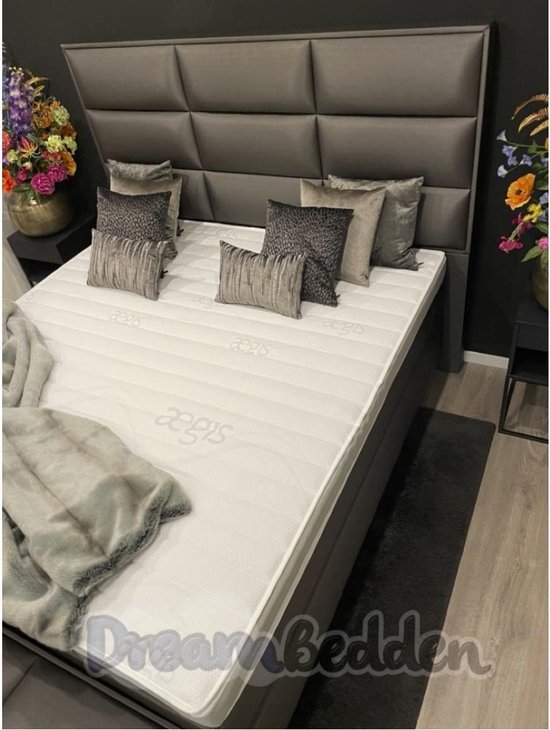 Grijze boxspring met hoog gestoffeerd hoofdbord, witte matras met Aegis-logo, grijze deken en sierkussens.