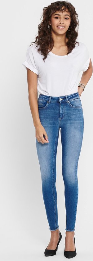 Vrouw met golvend bruin haar, draagt blauwe skinny jeans met rafelige zoom en witte top, zwarte pumps.