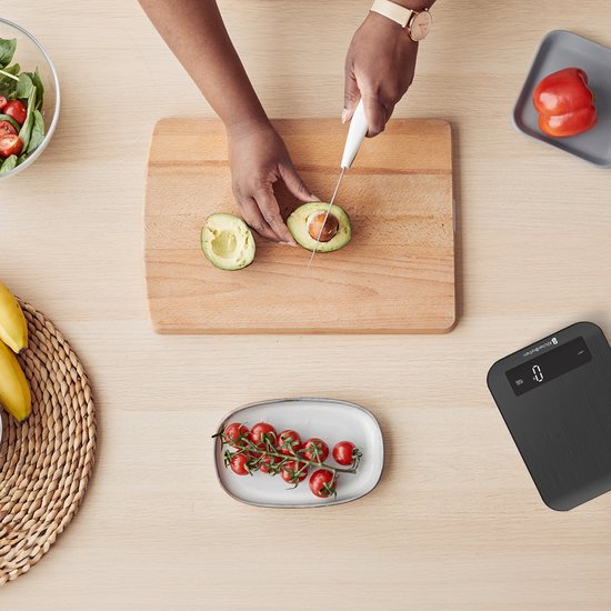 Balance de cuisine KitchenBrothers noire sur un plan de travail en bois, à côté d'un avocat coupé et de fruits.