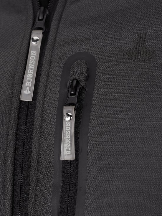 Close-up van de rits op de borst van een grijze softshell jas met Bjørnson tekst op de trekker.