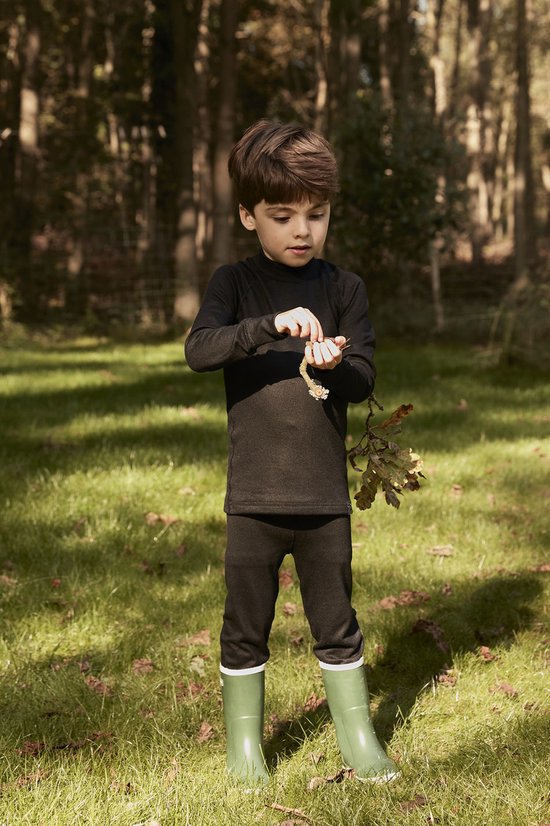 Jongen met zwarte thermokleding in een bosrijke omgeving, staand in groene laarzen.
