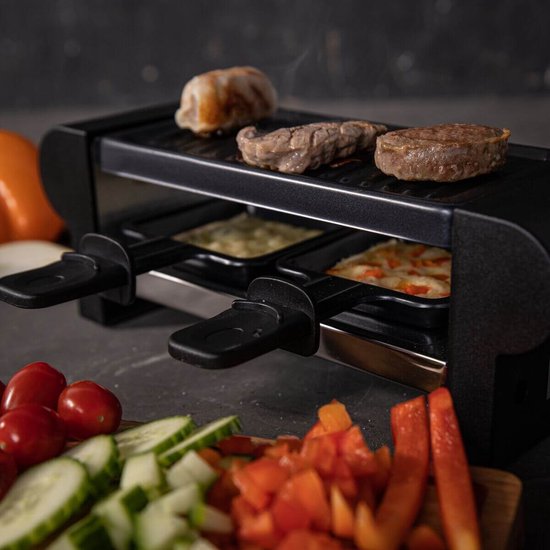 Raclette grill met gegrild vlees en gesmolten kaas in pannetjes, met gesneden groenten op de voorgrond.