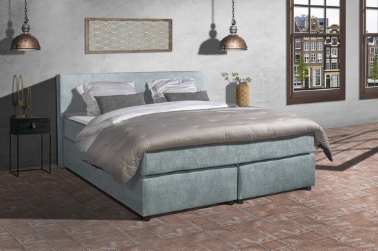 Een lichtblauwe boxspring met een gestoffeerd hoofdbord en nachtkastje, opgemaakt met grijs en wit beddengoed.