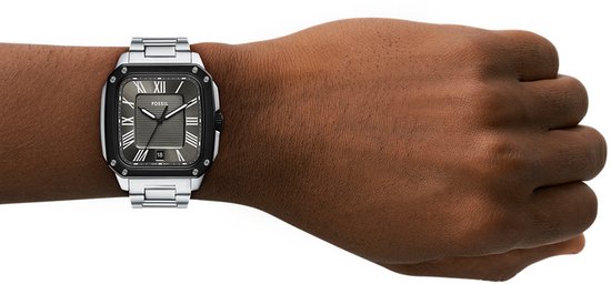 Zilverkleurig herenhorloge met vierkante wijzerplaat en zwarte rand, gedragen om een pols.
