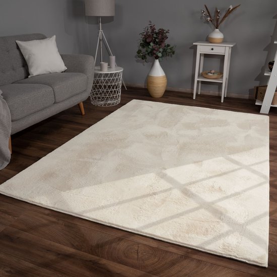 Tapis doux à poils longs crème avec des taches subtiles beiges, posé sur un parquet à côté d'un canapé gris.