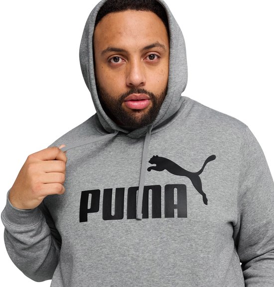 Close-up van een man in een grijze PUMA hoodie met zwart PUMA-logo op de borst. Hij trekt aan de capuchonkoord.