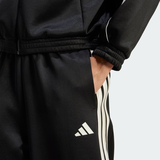 Detail van een zwarte trainingsbroek met beige 3-strepen en adidas logo op de zak.