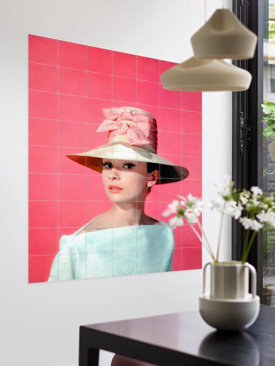 Audrey Hepburn portant un grand chapeau, un nœud rose et un haut pâle, sur fond rose, dans un intérieur moderne avec table d'appoint.