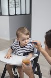Kind met streepjes T-shirt zit in een kinderstoel en eet met een blauw lepeltje. Een hand houdt een bakje met eten vast.