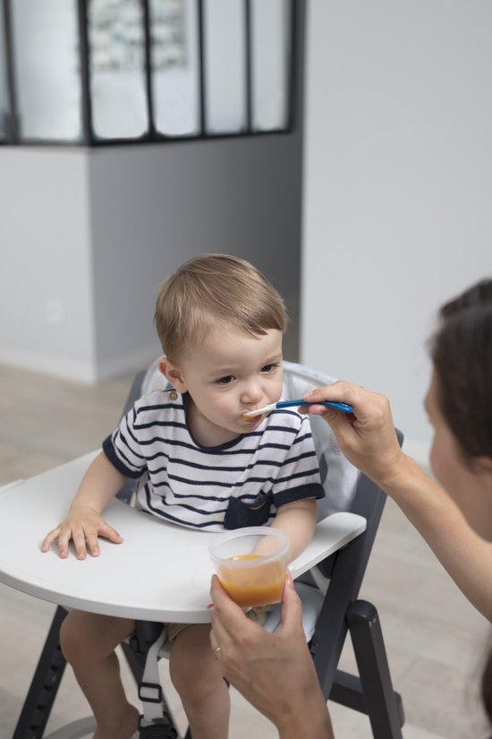 Kind met streepjes T-shirt zit in een kinderstoel en eet met een blauw lepeltje. Een hand houdt een bakje met eten vast.
