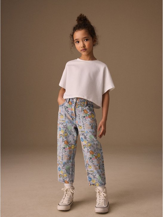 Meisje met opgestoken haar draagt een witte top en lichtblauwe, wijde jeans met all-over print van Smiley World-figuren.