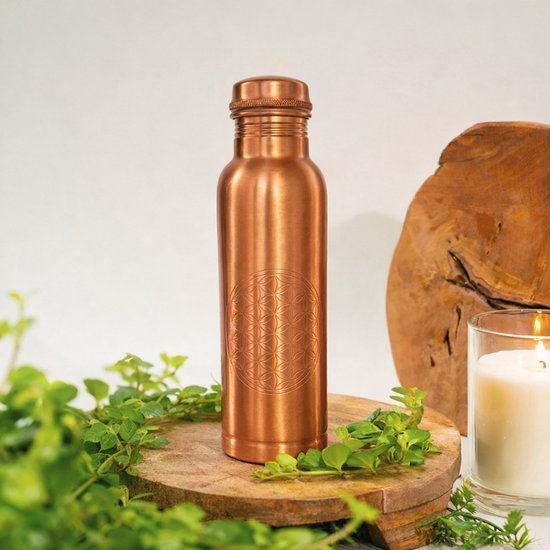 Koperen waterfles met 'Flower of Life' gravure, staand op een houten schijf, omringd door groen en een kaars.