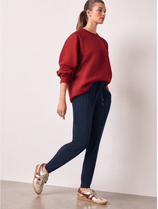 Model in een rode sweater en donkerblauwe joggingbroek met trekkoord, posed met gouden sneakers.