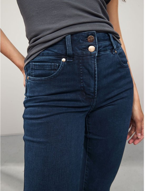 Model draagt een donkerblauwe spijkerbroek met bootcut pasvorm en slim-fit model. De broek heeft een hoge taille met dubbele knoopsluiting en gouden knopen.
