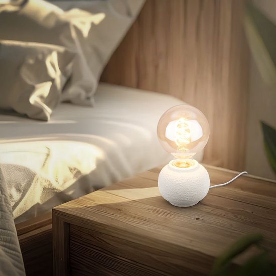 Lampe de table blanche à texture rugueuse, avec une grande ampoule chaude sur une table de chevet en bois près d'un lit.